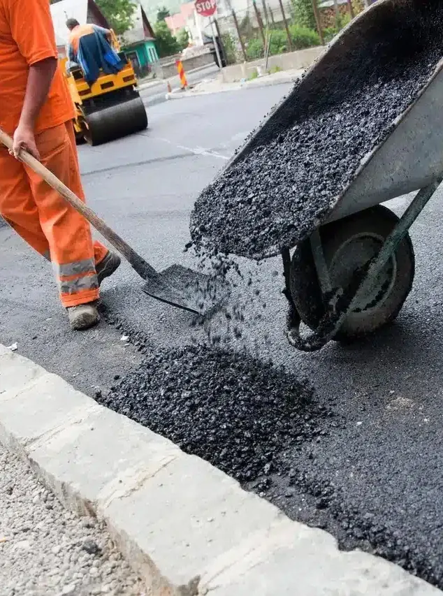 Munkás lapáttal teríti a talicskából kiöntött meleg aszfaltot a szegély mellett, henger a háttérben