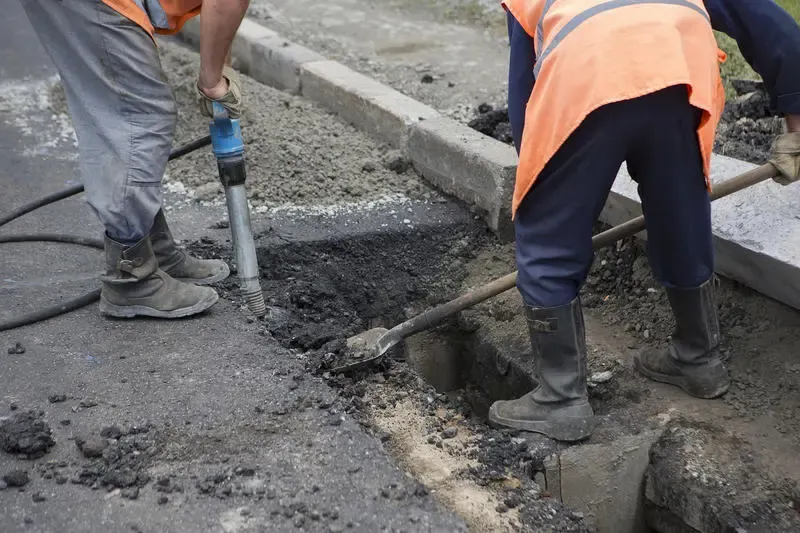 Két munkás légkalapáccsal és lapáttal bontja a sérült aszfaltot a szegélykő mellett