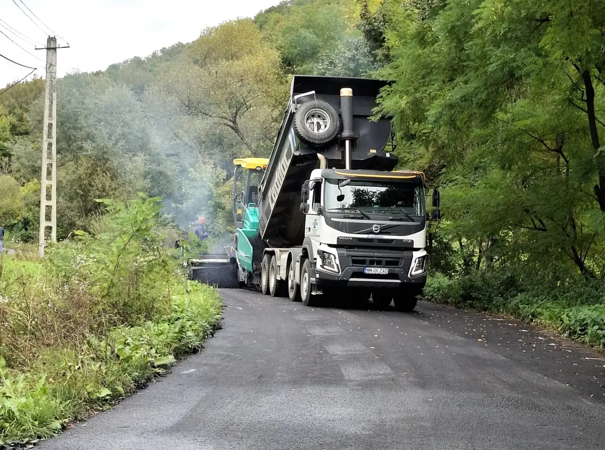 Volvo FMX billencs meleg aszfaltot ürít a Vögele terítőgépbe erdős, dombos útszakaszon
