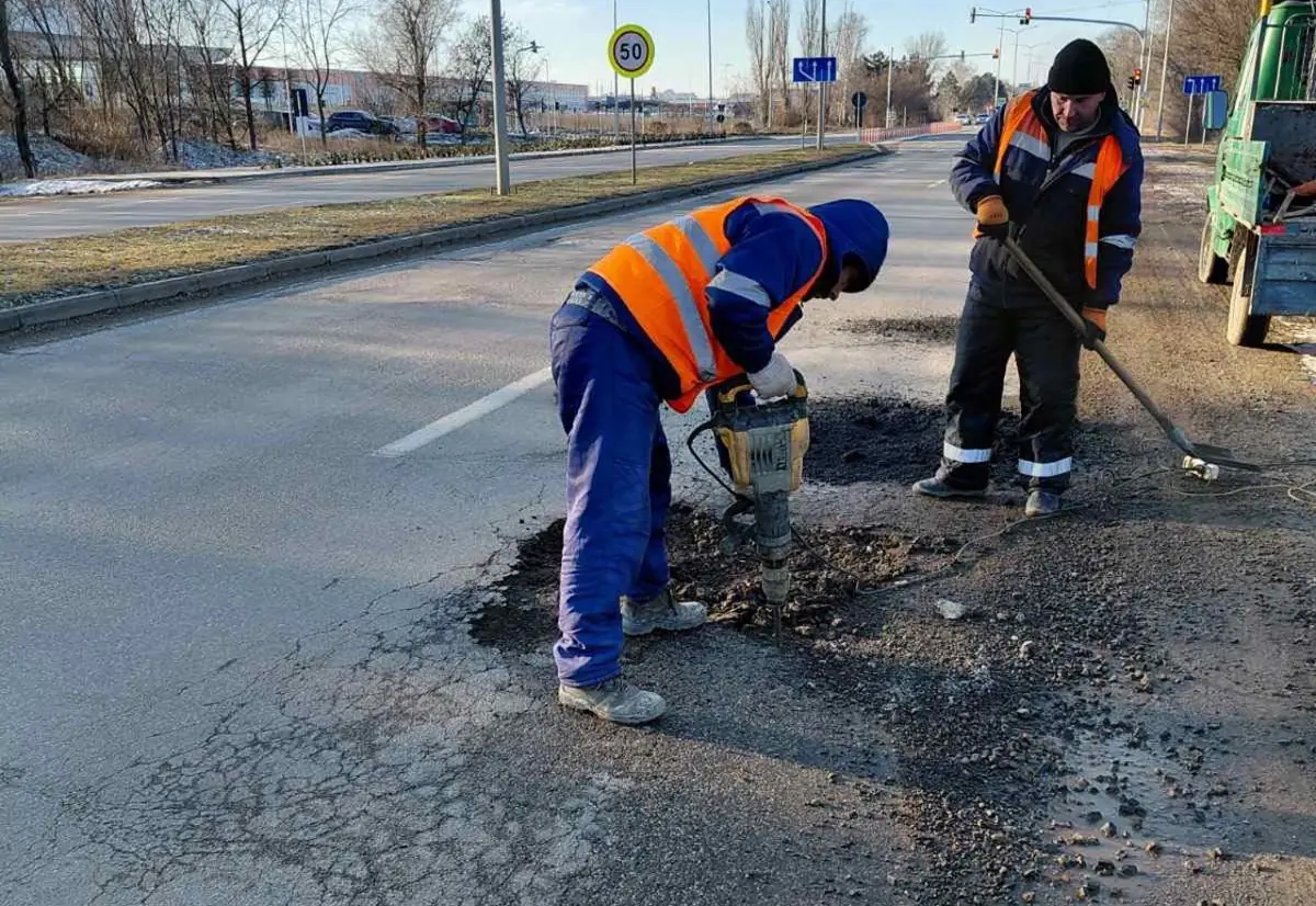 Munkás bontókalapáccsal bontja a kátyú szélét havas, téli városi úton, társa lapáttal tisztít