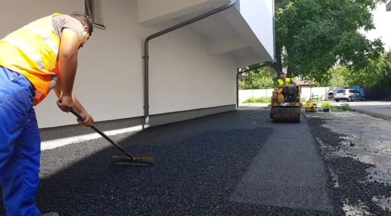 Munkás gereblyével teríti a friss aszfaltot egy fehér épület fala mellett, a háttérben kis henger dolgozik