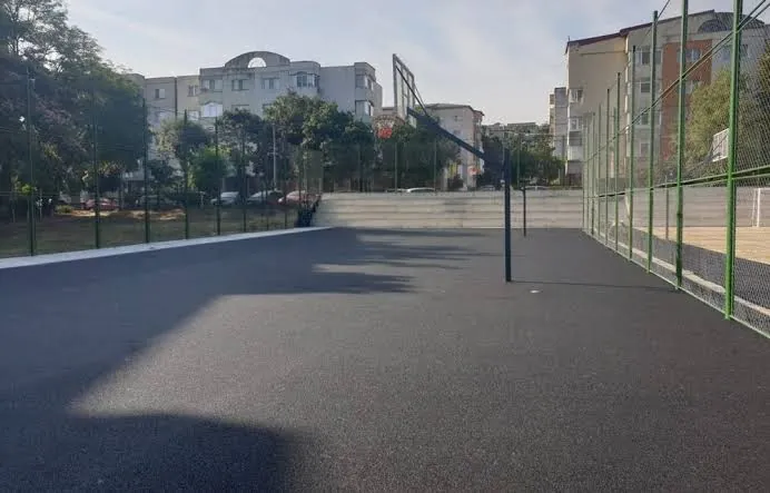 Elkészült, kerítéssel körbevett aszfaltozott sportpálya kosárlabda-palánkkal, lakóházak között