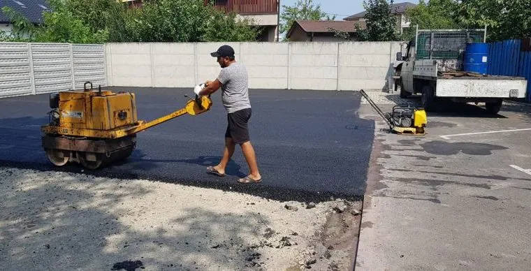 Munkás vezetett hengerrel tömöríti a friss aszfaltot betonkerítéssel körülvett telephelyi udvaron, lapvibrátor a háttérben